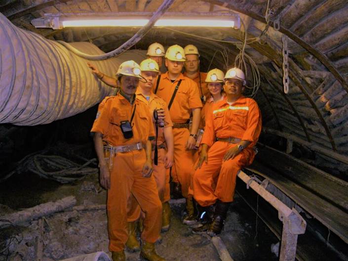 Seven people in orange coveralls and hard hats standing together in an arched underground coal mine tunnel, with a ventilation duct along the left wall and a lamp overhead.